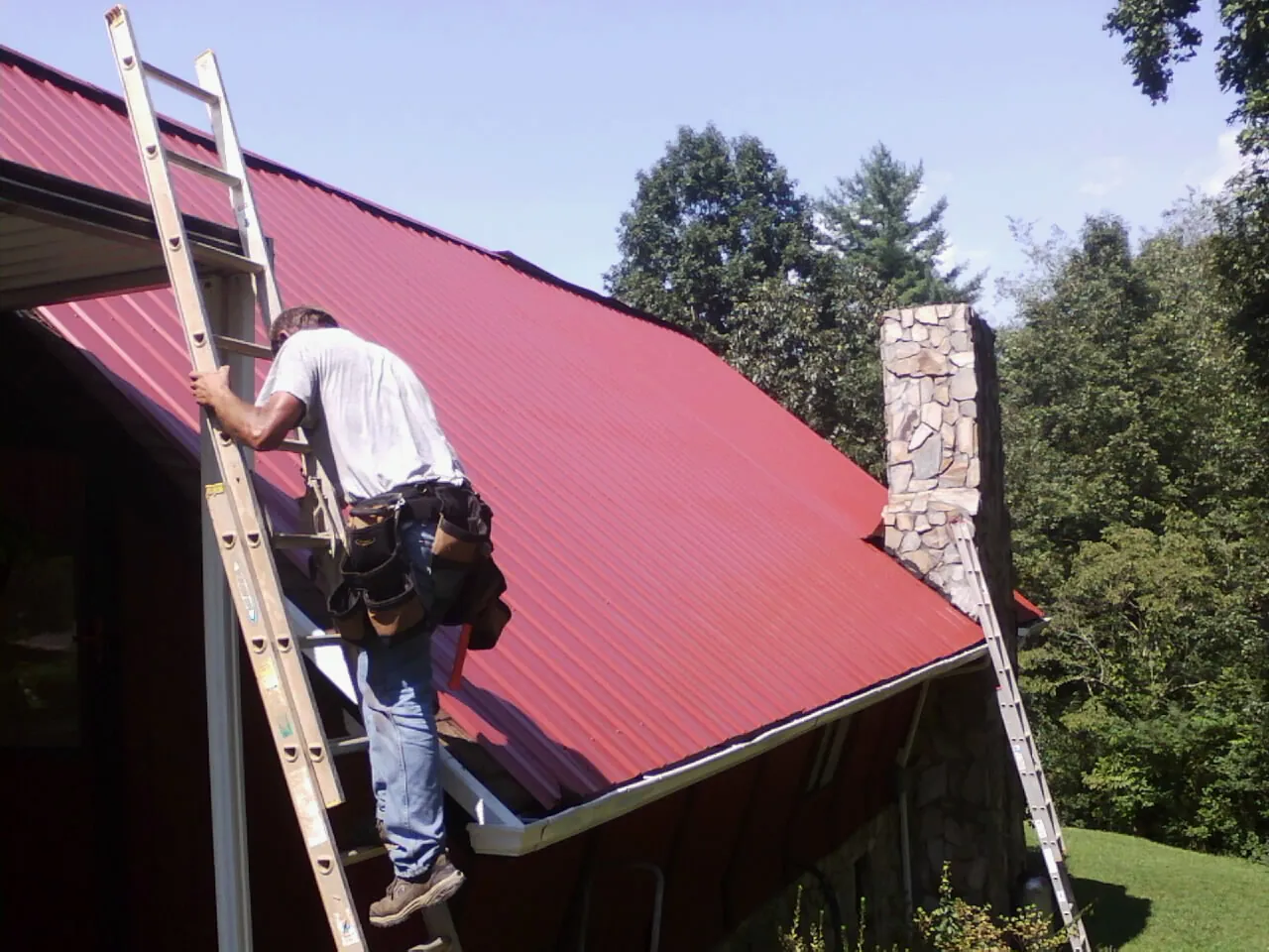 Roofer on a ladder beside a red metal roof and stone chimney — Soffit & Fascia Repair by Roofing Brothers in Walland